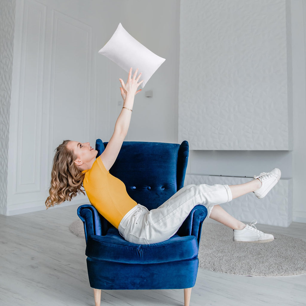 Woman in yellow top and white pants playfully tossing a white pillow while sitting in a blue armchair in a bright room.
