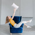 Woman in yellow top and white pants playfully tossing a white pillow while sitting in a blue armchair in a bright room.