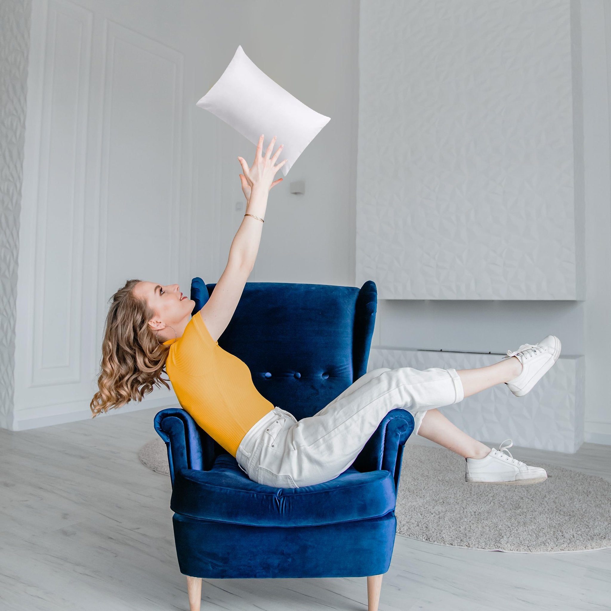 Woman in yellow top and white pants playfully tossing a white pillow while sitting in a blue armchair in a bright room.