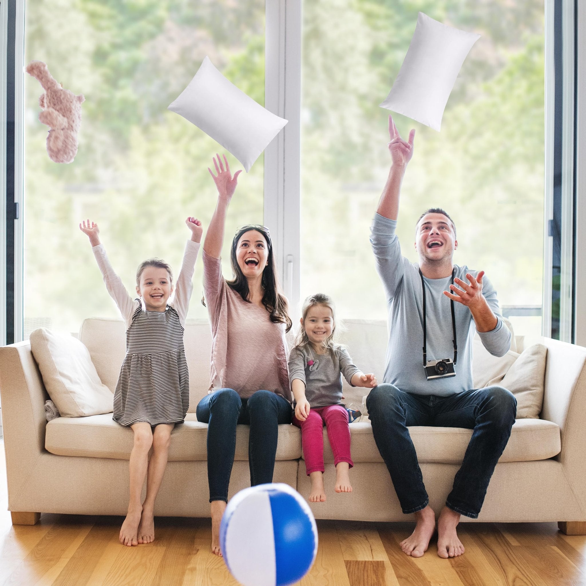 Family of four playing with white pillow inserts and a stuffed animal in a living room with a large window