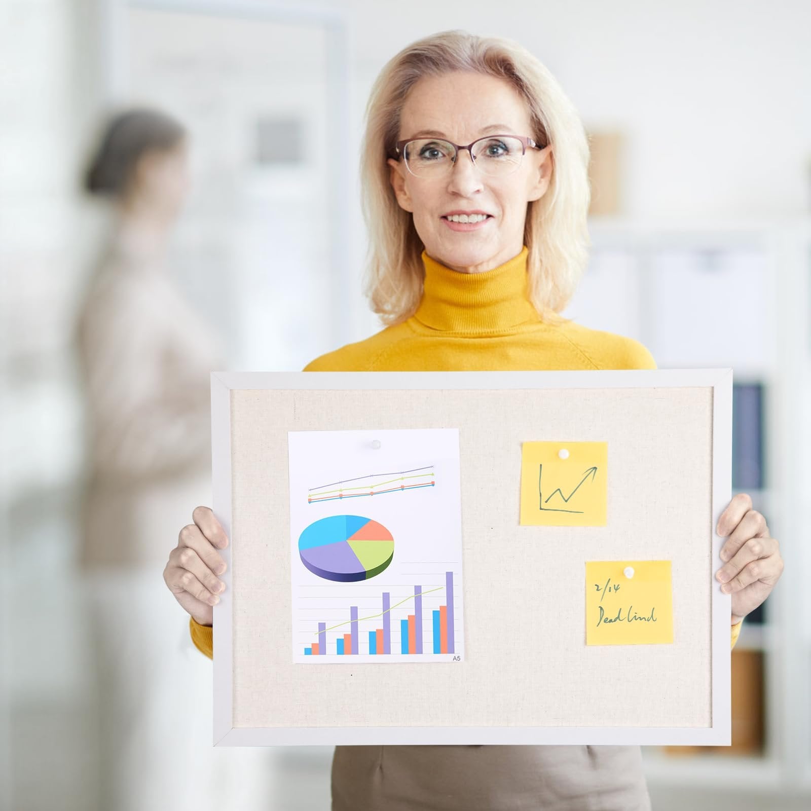 Woman holding a linen bulletin board with a white wood frame, displaying charts and sticky notes.