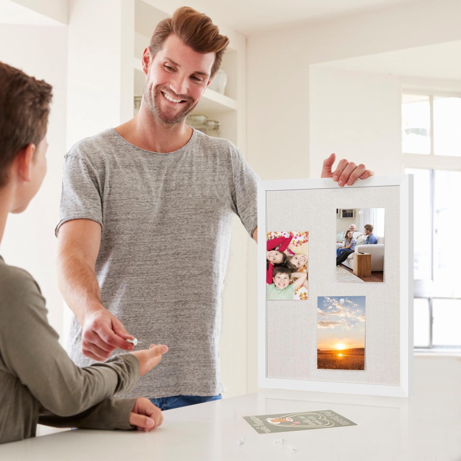 Man showing a linen bulletin board with white wood frame to a child, with photos pinned to it.