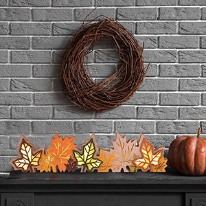 Wooden maple leaf block sign with LED lights and a grapevine wreath above, set on a mantelpiece with a pumpkin to the right.
