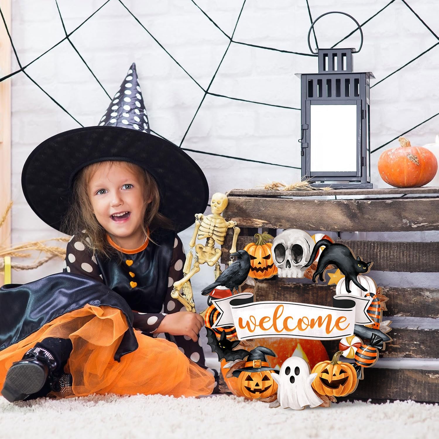 Girl in witch costume sitting next to Halloween wreath with welcome sign, skeleton, and lantern