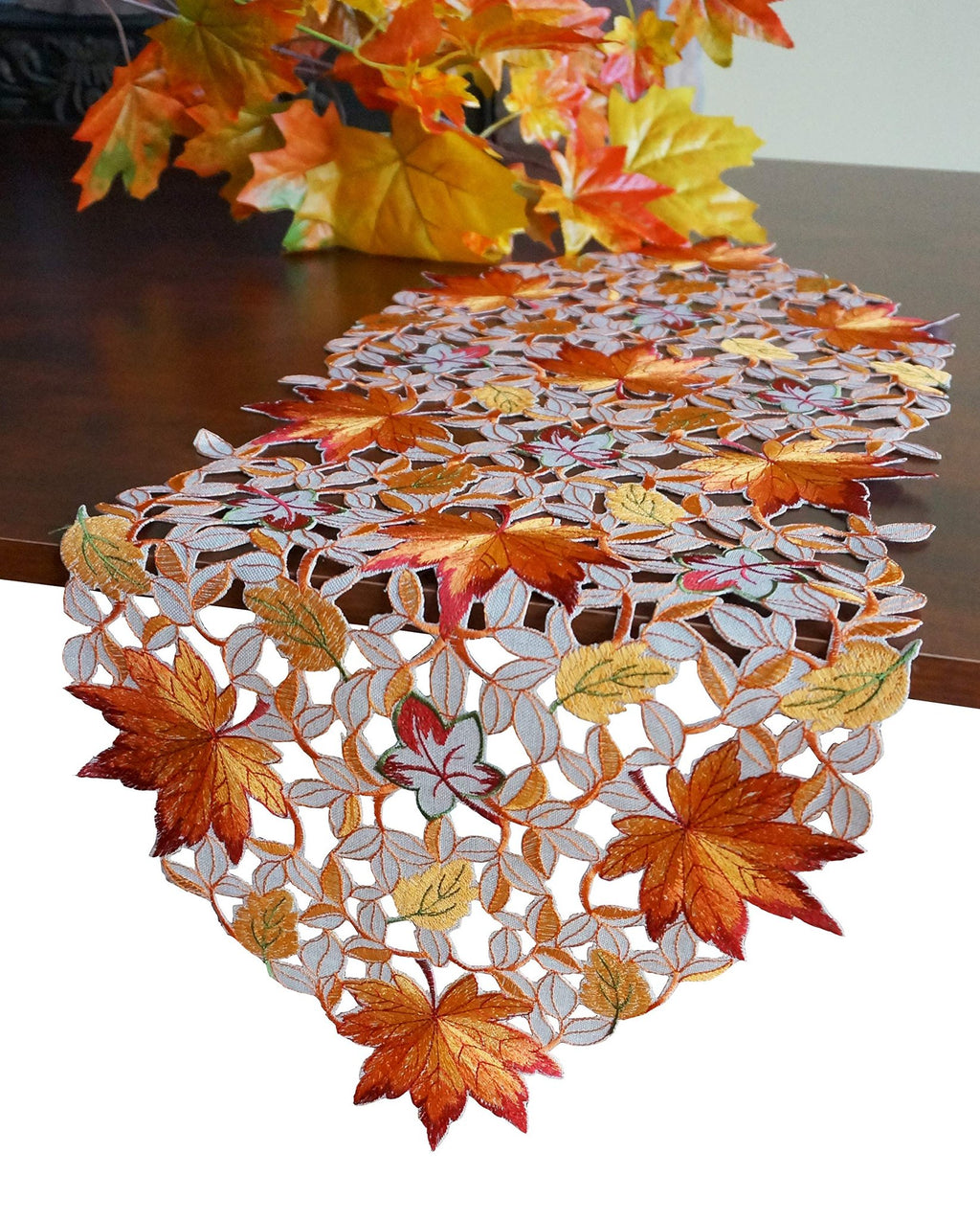 Fall table runner with embroidered maple leaves and cutwork design on a wooden table with autumn foliage in the background.