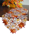 Fall table runner with embroidered maple leaves and cutwork design on a wooden table with autumn foliage in the background.