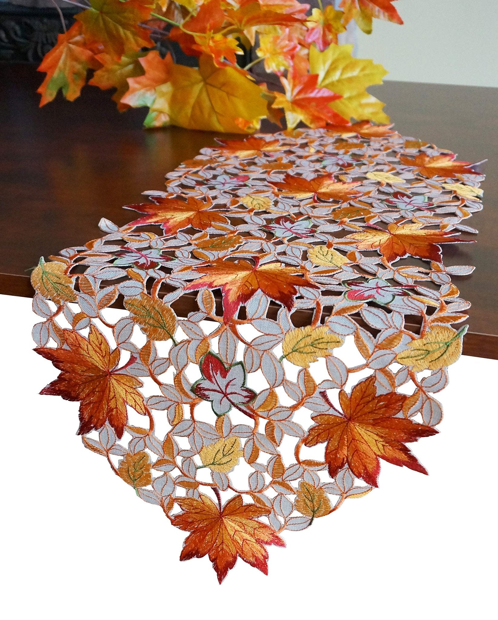 Fall table runner with embroidered maple leaves and cutwork design on a wooden table with autumn foliage in the background.