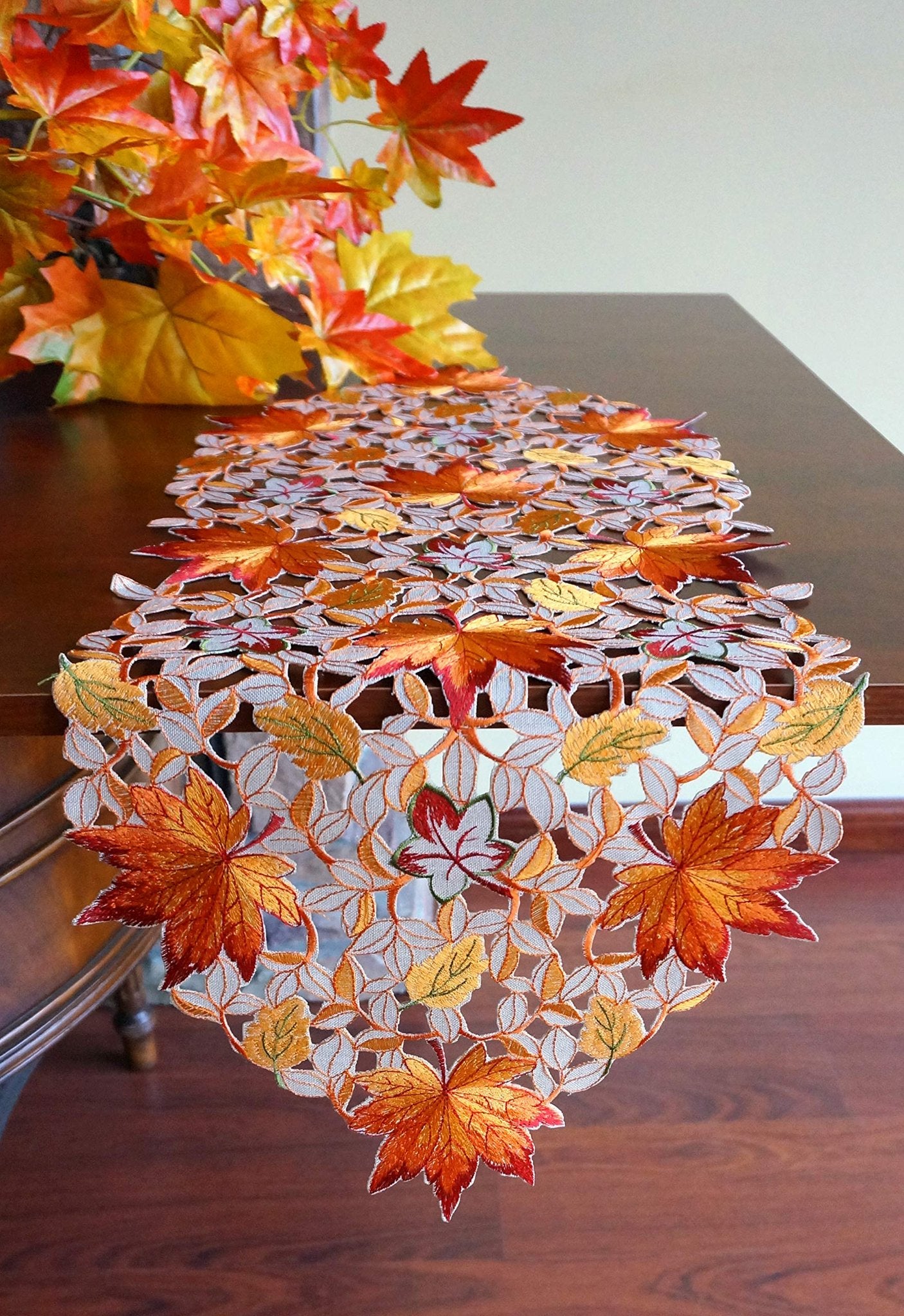 Fall table runner with embroidered maple leaves and cutwork pattern on a wooden table, with autumn foliage in the background.