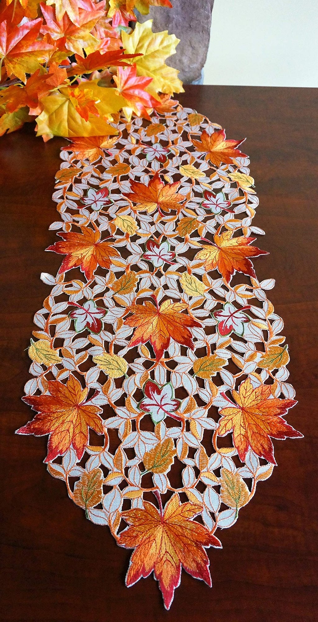 Fall table runner with embroidered maple leaves and cutwork pattern, displayed on a wooden table with autumn leaves.