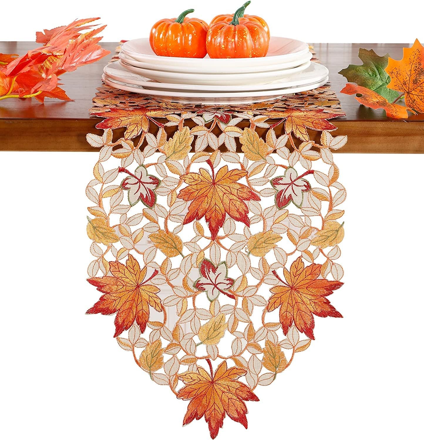Fall table runner with embroidered pumpkins, maple leaves, and bats on a wooden table with plates and decorative leaves.
