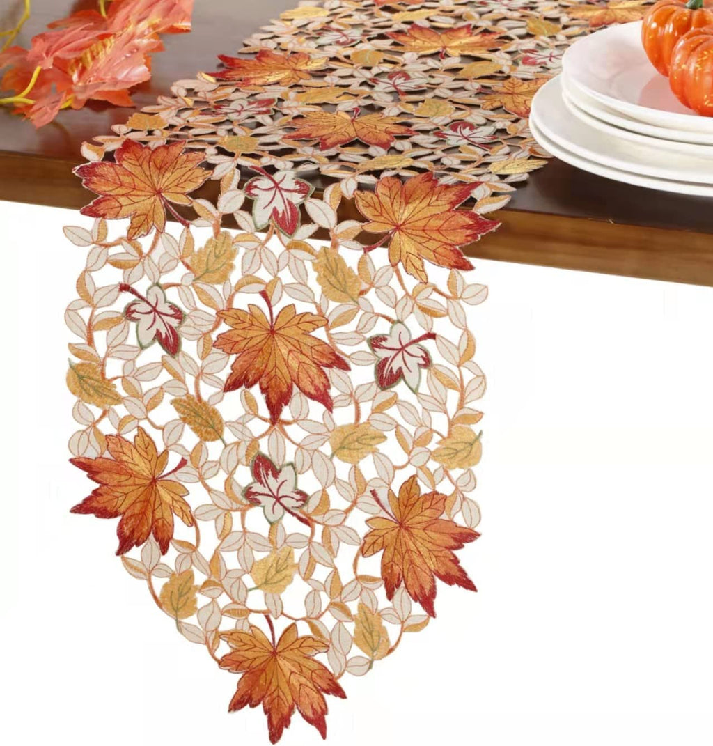 Fall table runner with embroidered autumn leaves and cutwork design, draped on a wooden table with decorative pumpkins and plates.