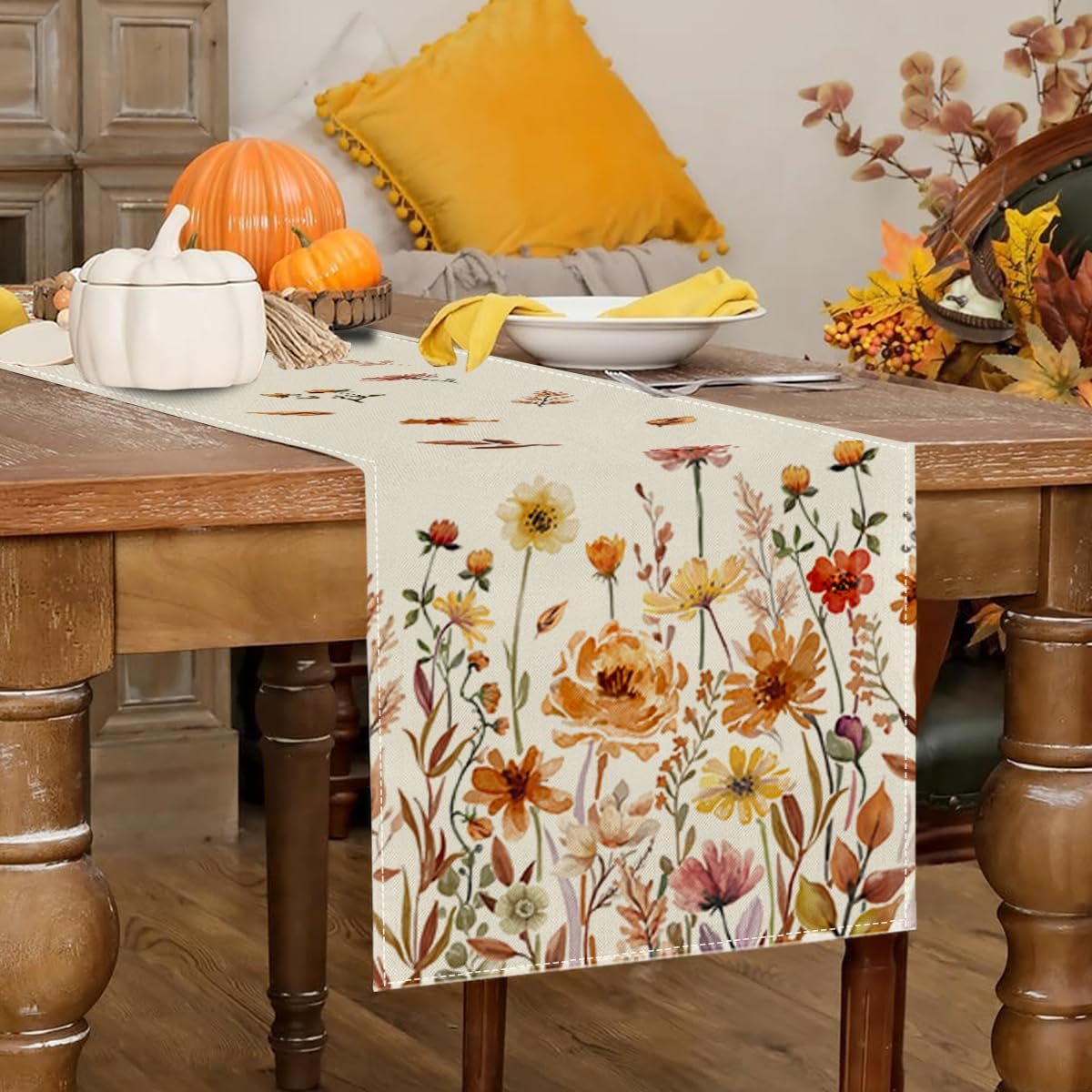 Fall floral table runner with autumn leaves and flowers displayed on a wooden dining table with pumpkins and a yellow pillow.