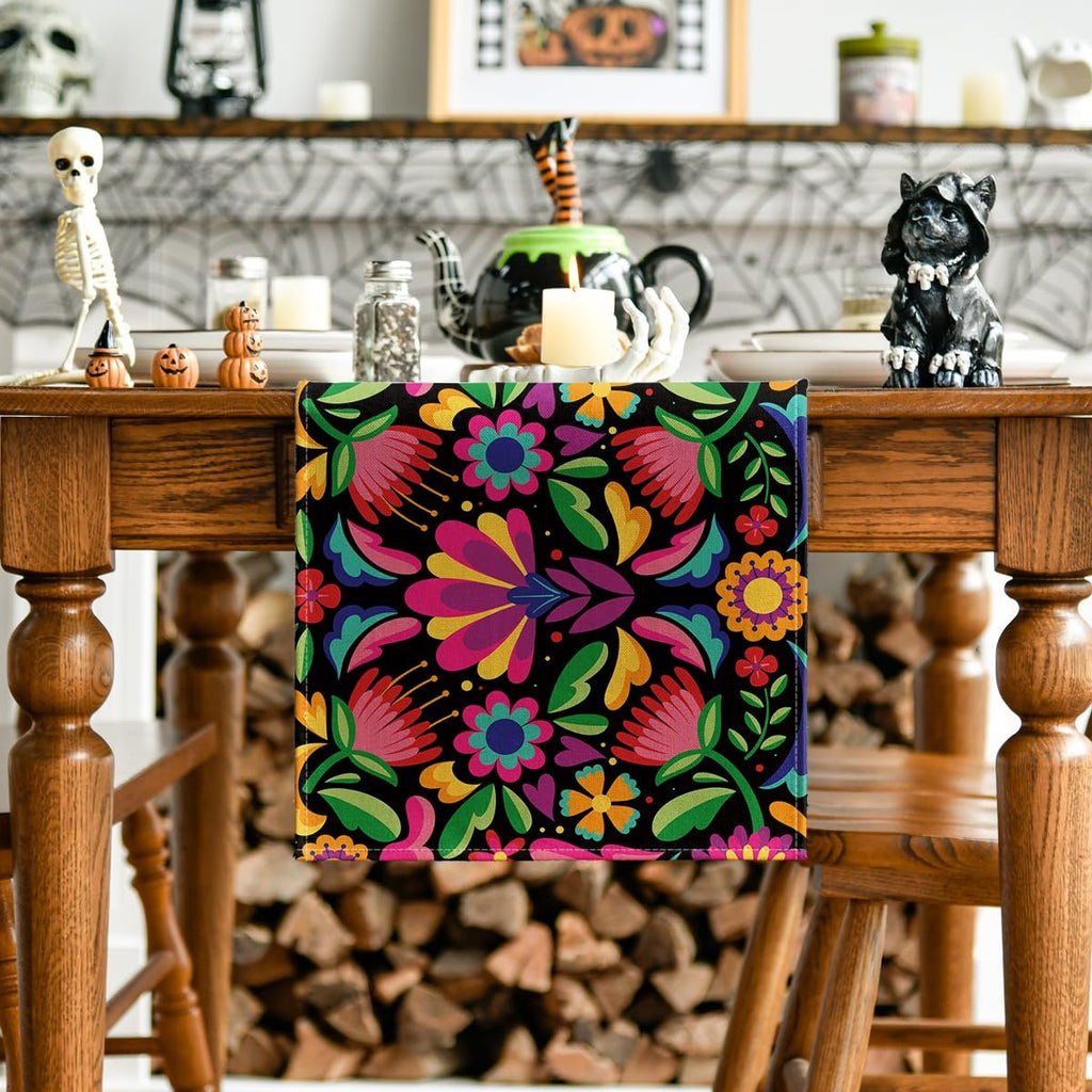 Floral serape table runner with colorful embroidered flowers on a black background, draped over a wooden table with Halloween decorations.