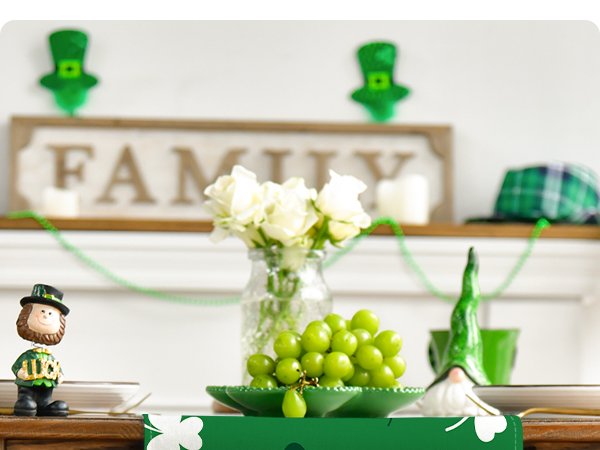 Green St. Patrick's Day table runner with shamrocks and leprechaun decor on a table with grapes and flowers.