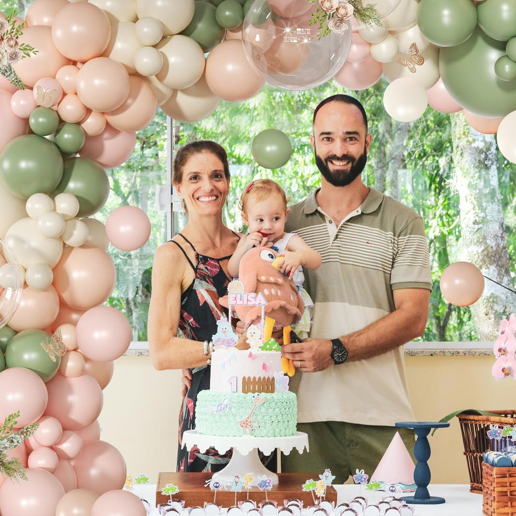 Family celebrating a baby's first birthday with a pink and sage green balloon arch and cake.