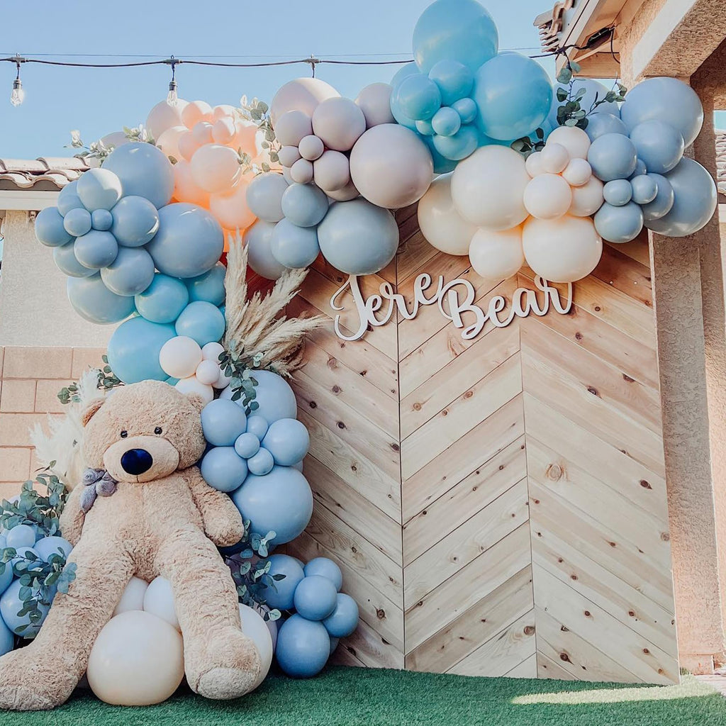 Balloon arch with dusty light blue, baby blue, and peach balloons, a large teddy bear, and wooden wall backdrop.