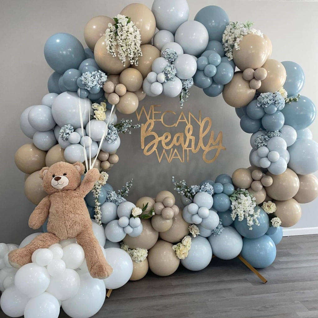 Balloon arch with a teddy bear and 'We Can Bearly Wait' sign, featuring dusty blue, light blue, and tan balloons with white flowers.