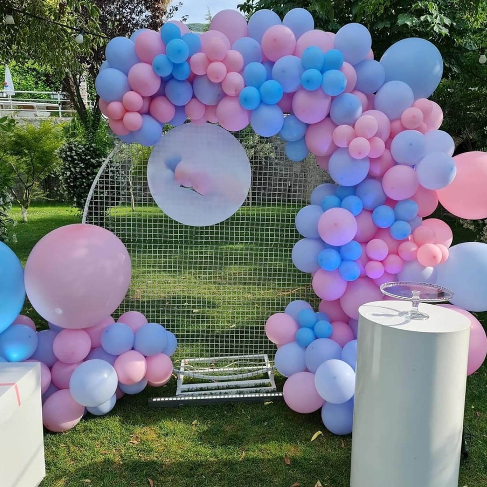 Pink and blue balloon garland arch setup outdoors with a white mesh backdrop and a cake stand
