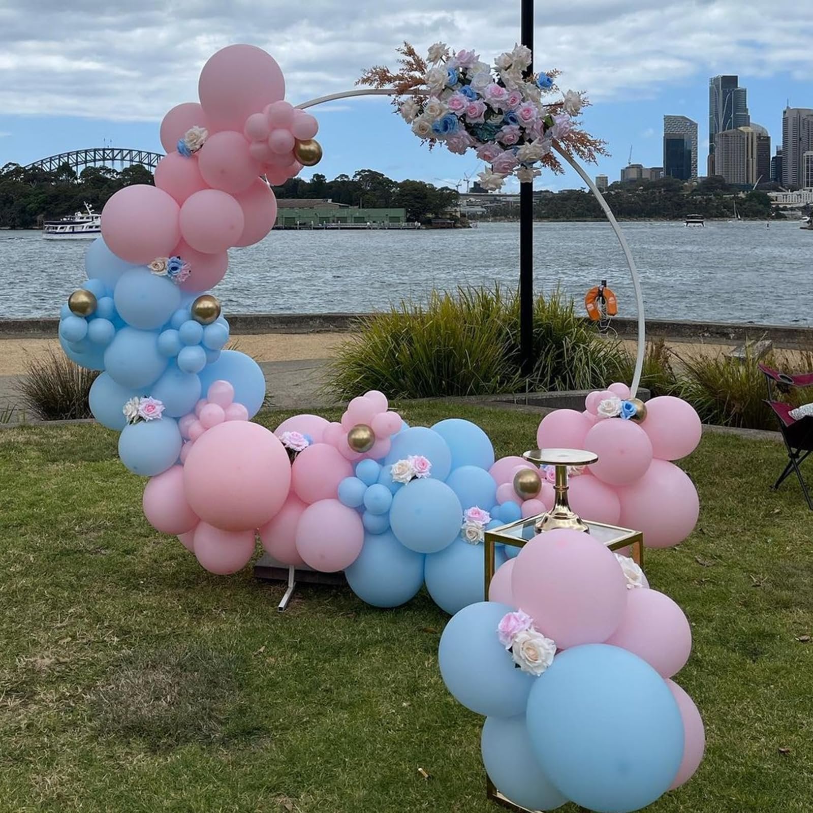 Pink and blue balloon garland arch with floral accents and gold spheres, set outdoors by a body of water with city skyline in background.