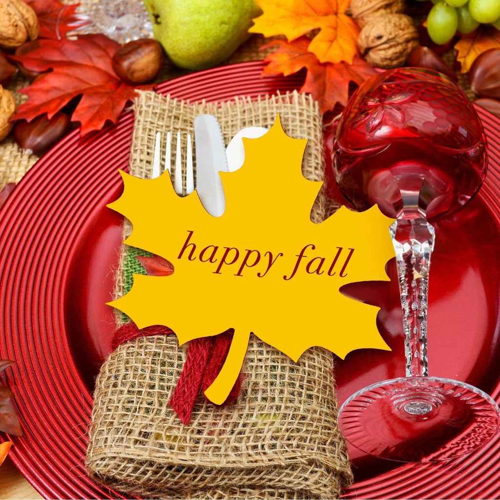 Yellow maple leaf cutout with 'happy fall' text on a Thanksgiving table setting with red plates and autumn decor.