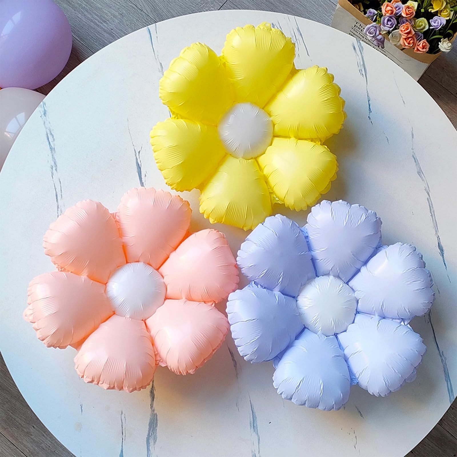 Three pastel flower-shaped foil balloons in yellow, pink, and lavender on a white table with a bouquet of flowers in the background.