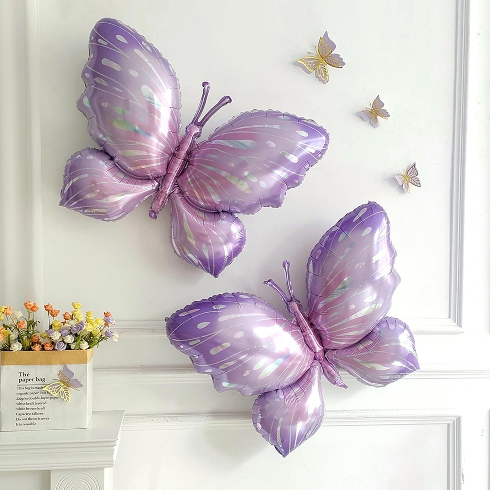Two large purple butterfly foil balloons attached to a white wall, with smaller gold butterfly decorations scattered around. A vase of flowers sits on a shelf below.