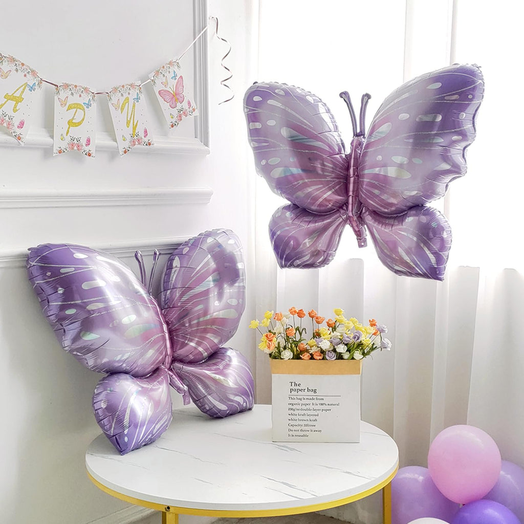 Two large purple butterfly foil balloons and a banner with letters spelling 'HAPPY' on a table with flowers and balloons.