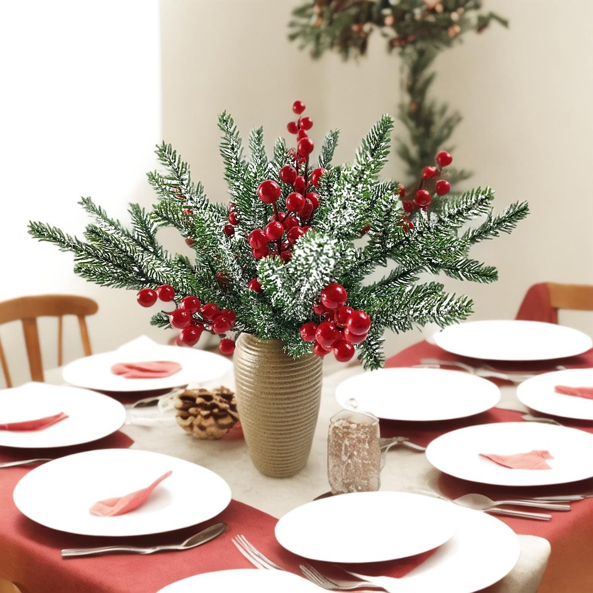 Frosted artificial pine branches with red berries in a vase on a table set for a meal.