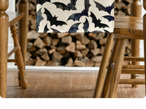 Black bats pattern table runner draped over a wooden table between chairs, with a stack of firewood in the background.