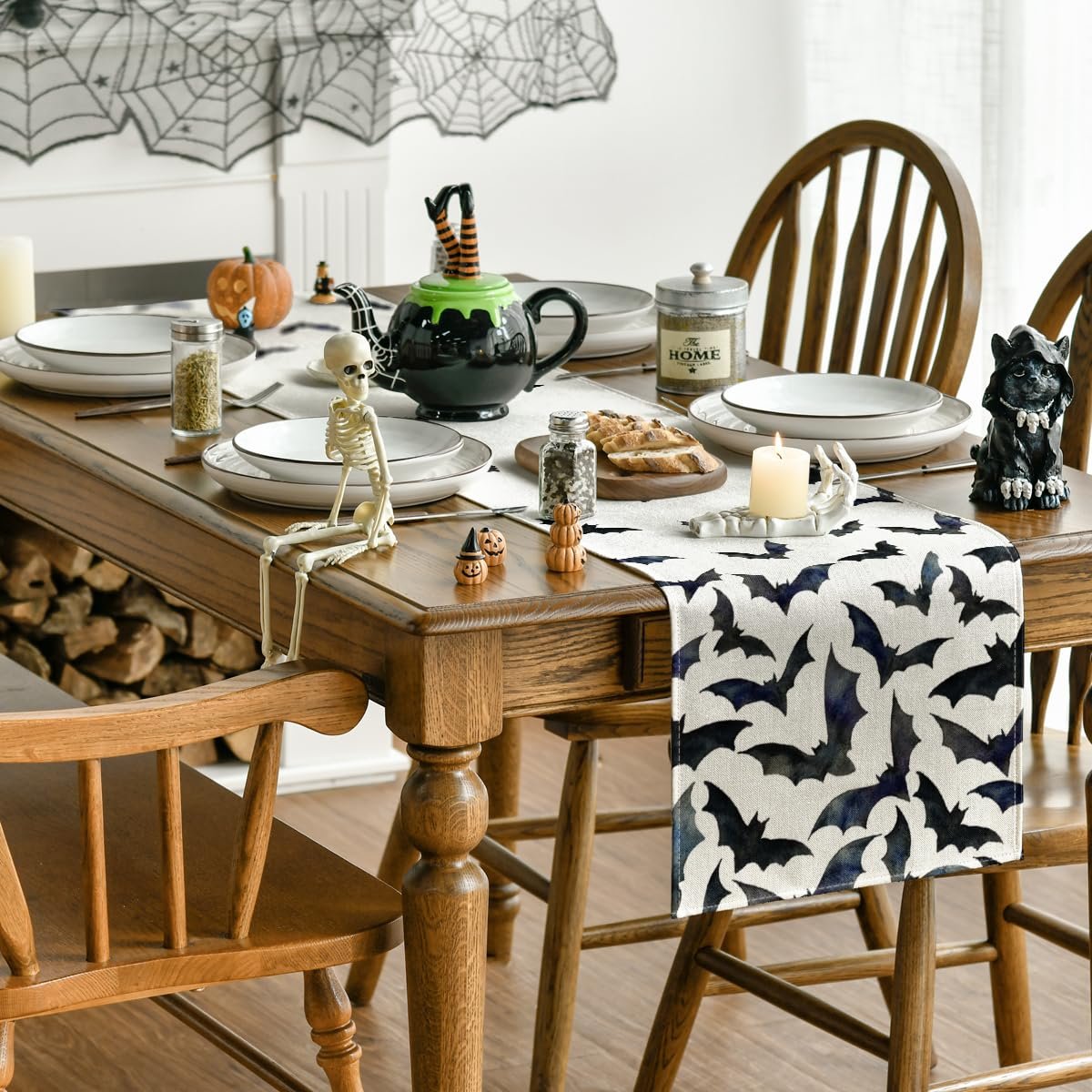 Halloween table runner with black bats pattern on a wooden dining table set for a meal, featuring skeleton and witch teapot decorations.