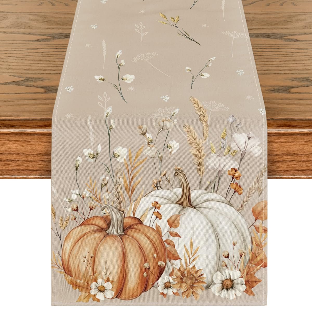 Fall table runner with pumpkins, white flowers, and wheat stalks on a beige background, draped over a wooden table.