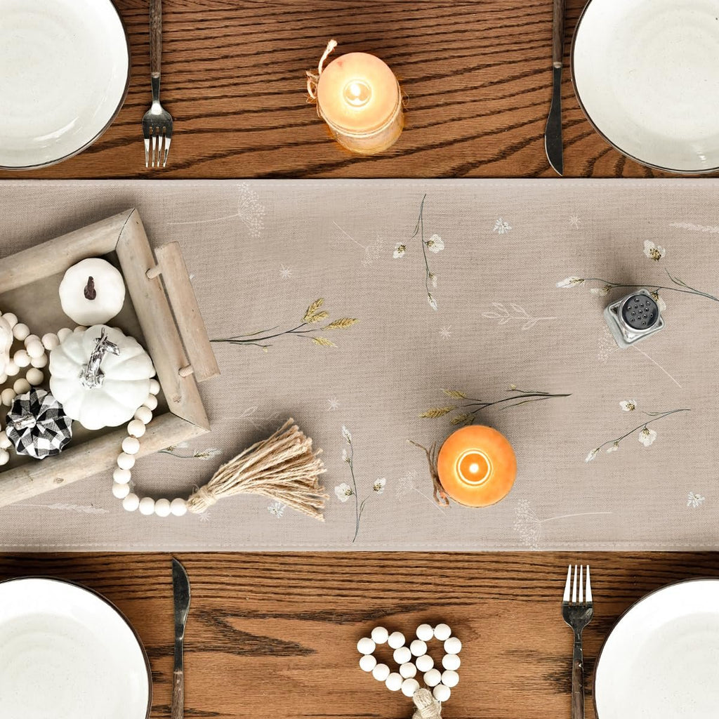 Fall table runner with pumpkins, leaves, and flowers print, set on a wooden table with place settings and candles.