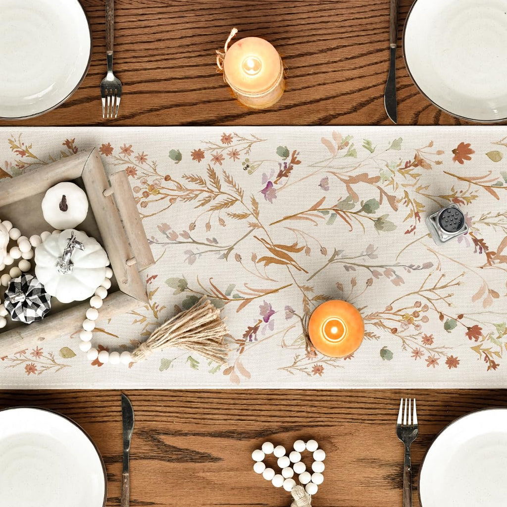 Table setting with a floral leaves fall table runner, white plates, cutlery, candles, and decorative pumpkins.