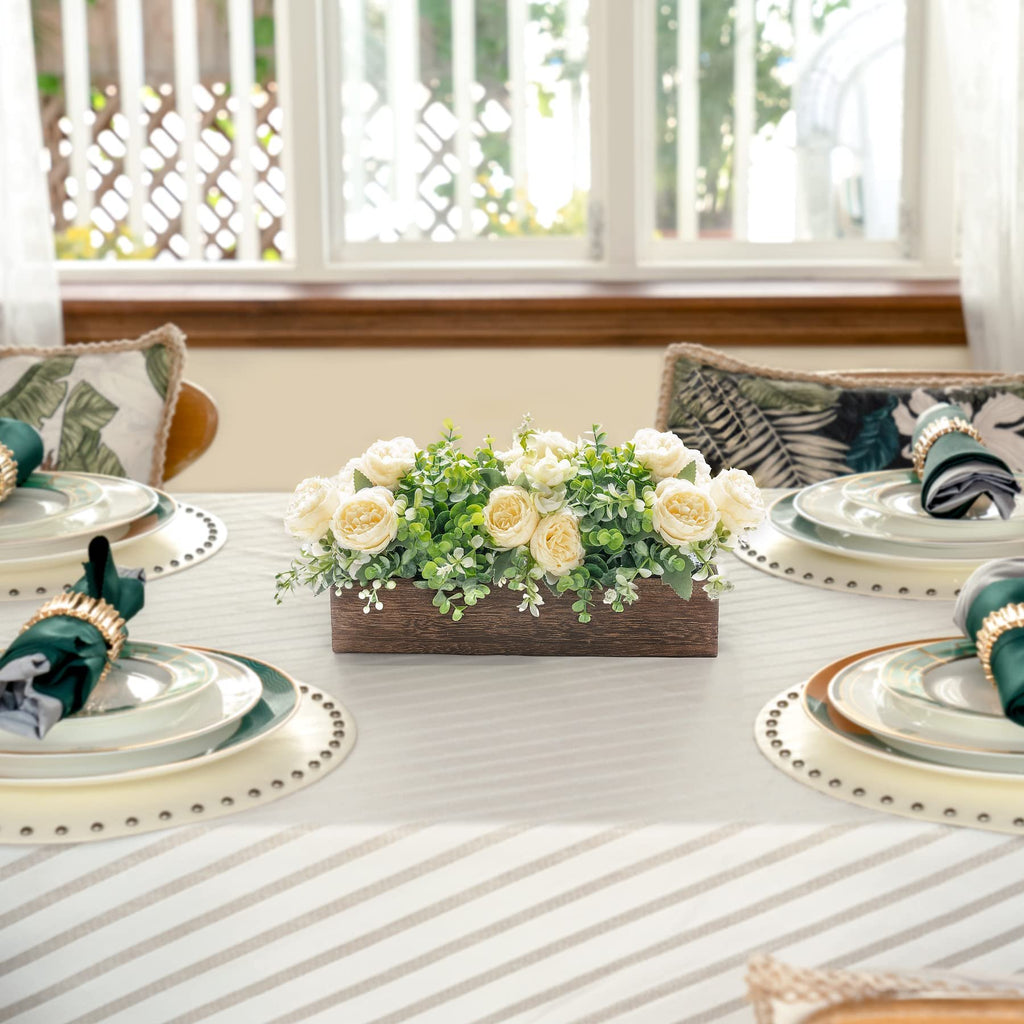 Wooden centerpiece box with artificial yellow roses and green eucalyptus on a dining table set for a meal.