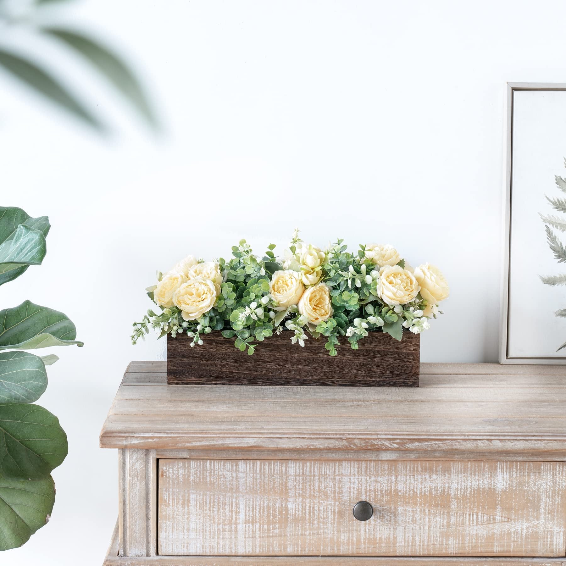 Wooden centerpiece box with yellow roses and eucalyptus on a rustic wooden table, with a large leaf plant and framed art in the background.