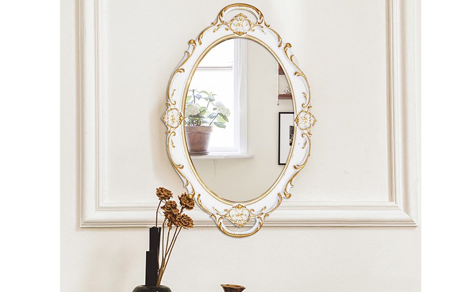 Oval antique decorative wall mirror with white and gold ornate frame, reflecting a window with plants.
