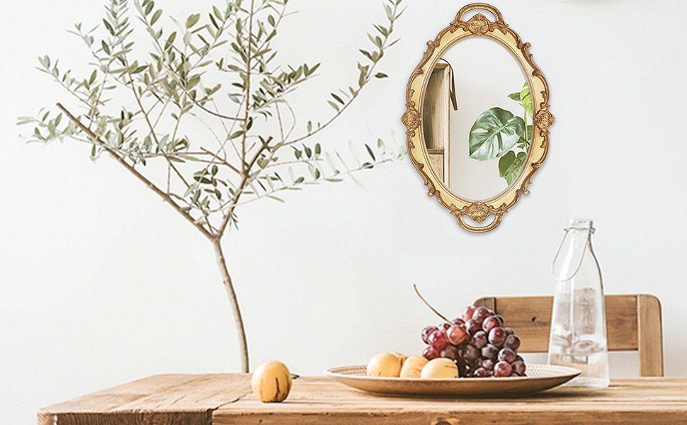 Oval antique decorative wall mirror in gold finish hanging on a white wall, with a table setting in the foreground.