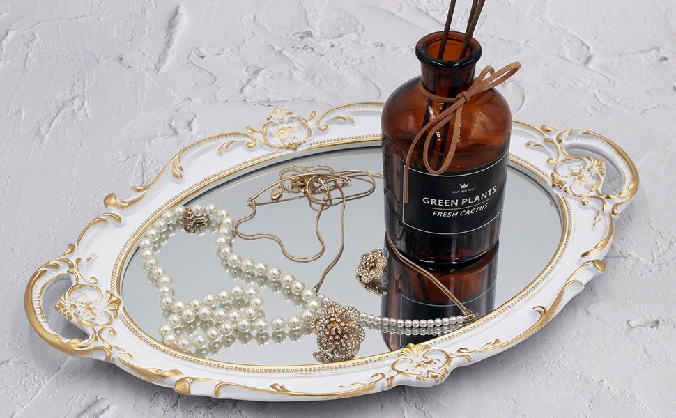 White and gold ornate oval decorative tray with mirrored surface holding pearl necklace, earrings, and a decorative bottle.