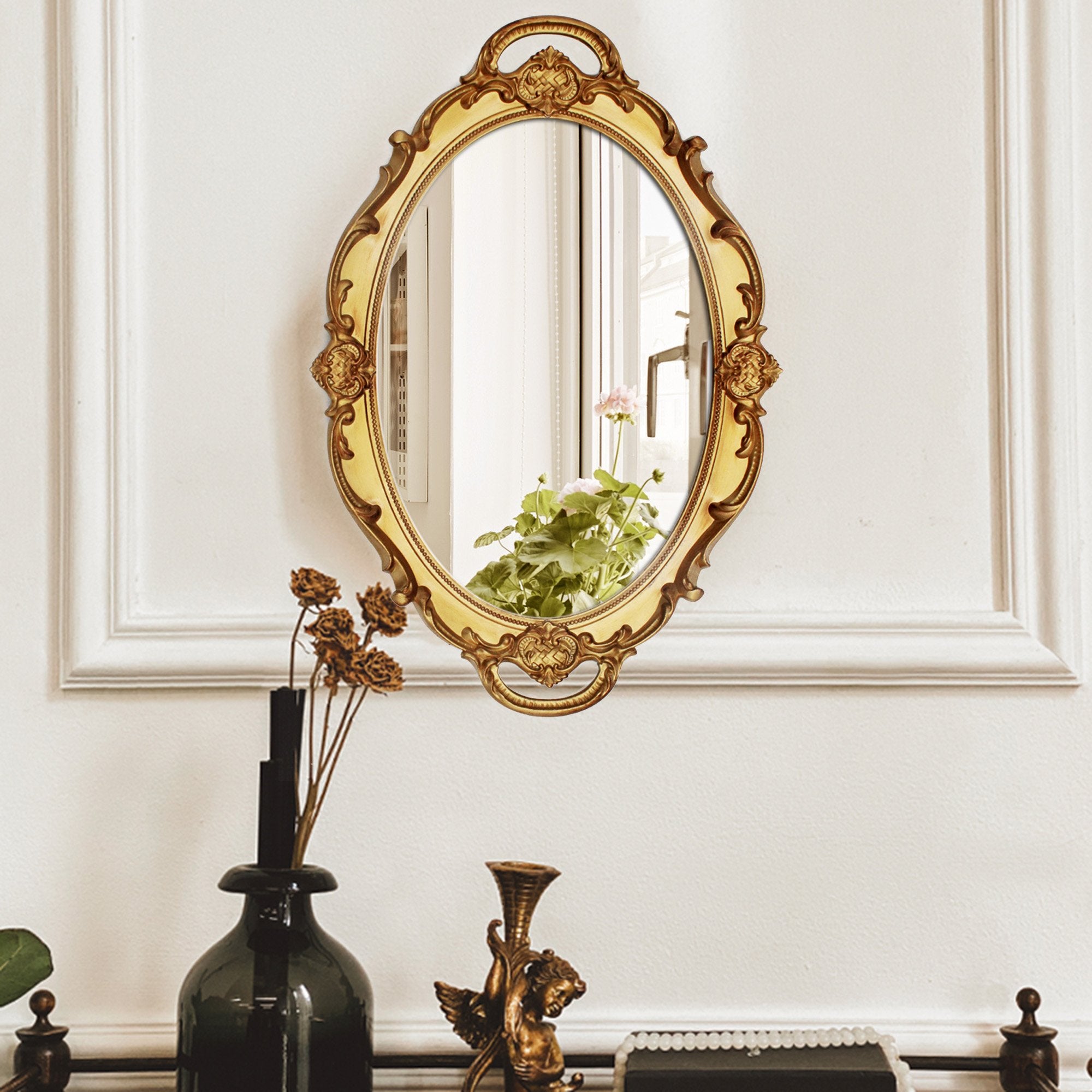 Ornate oval antique decorative wall mirror with gold frame hanging on a white wall above a shelf with decorative items.