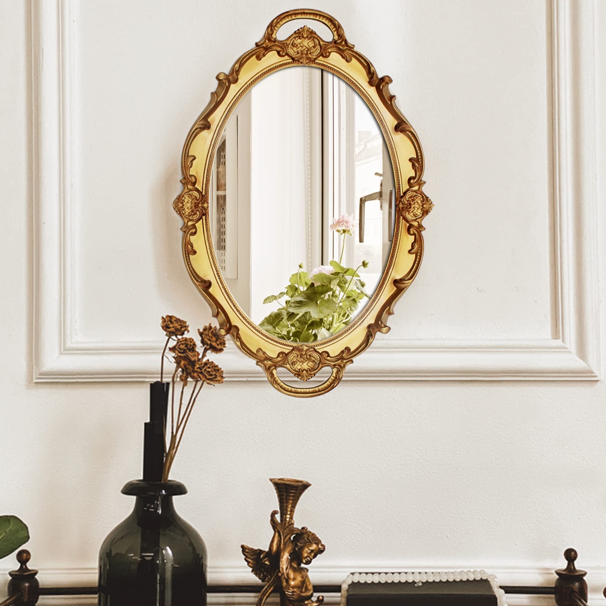 Oval antique decorative wall mirror with ornate gold frame hanging on a white wall, reflecting plants and window.