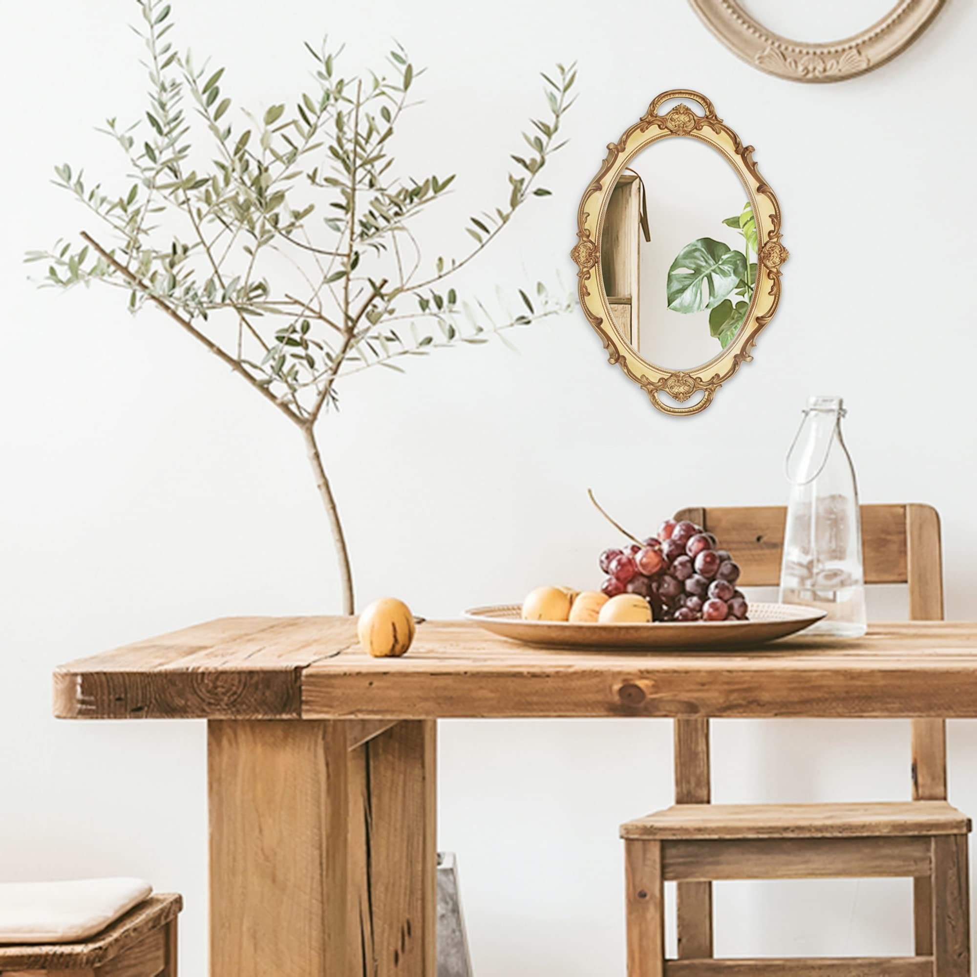Oval antique decorative wall mirror with gold ornate frame hanging above a wooden table with fruit and a bottle of water.
