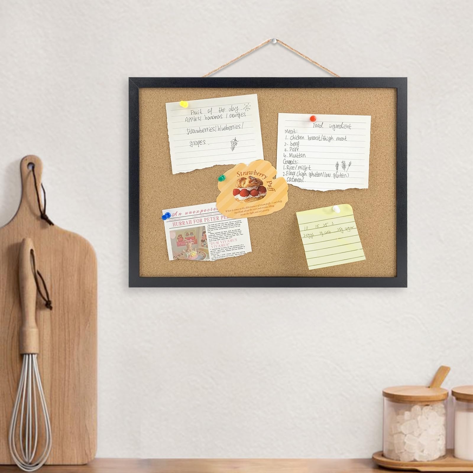 Black cork bulletin board with oak wood finish frame, hung on a wall with notes and a strawberry pastry sticker pinned to it.