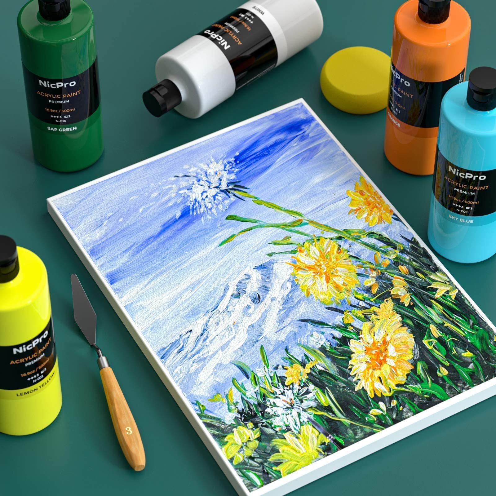 Acrylic paint bottles and a painting of a mountain with yellow flowers, arranged on a dark green surface.