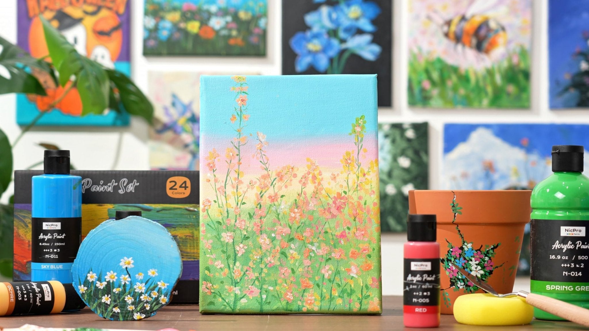 Acrylic paint bottles and a painted canvas with flowers, set against a backdrop of various paintings.