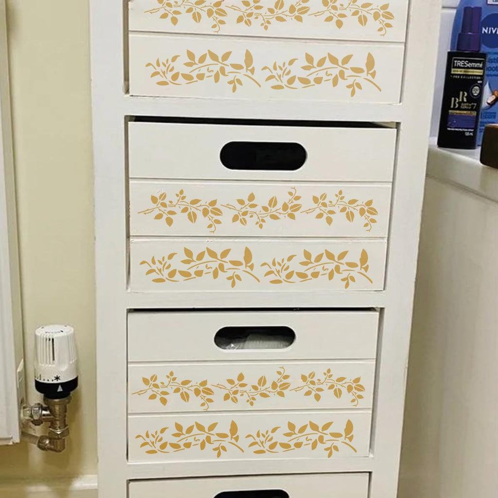 White wooden drawer unit with gold vine leaf stencils applied to drawer fronts. Includes a radiator and toiletries on a shelf.