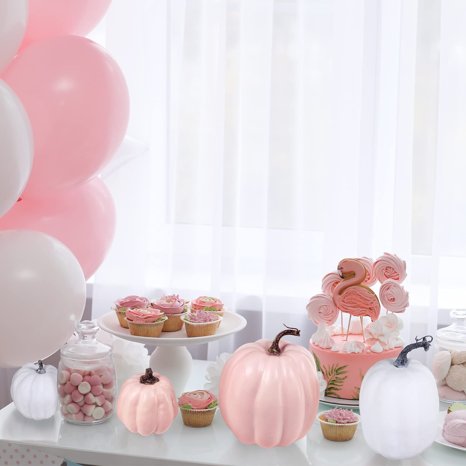 Assorted pink and white faux pumpkins arranged on a table with cupcakes and balloons for a party setting.