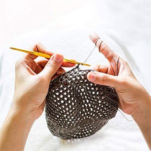 Hands crocheting with a gold aluminum hook and dark gray yarn on a white surface
