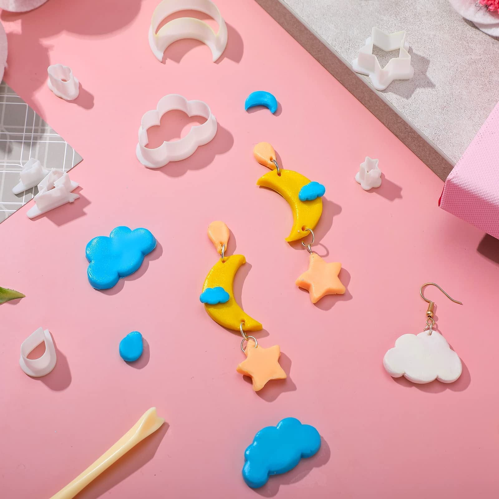 Polymer clay cutters and finished jewelry pieces in cloud, moon, star, and raindrop shapes on a pink background.