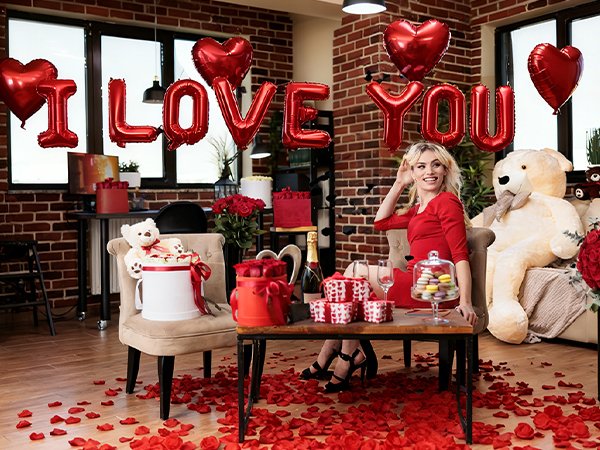 Woman in red dress sitting with 'I LOVE YOU' balloons and rose petals for Valentine's Day celebration