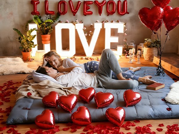 Couple in bed surrounded by red heart balloons and rose petals, with 'I LOVE YOU' balloons overhead.